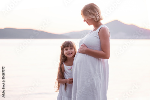 Close-up portrait of a little girl hugging pregnant mother. A daughter in a white sundress strokes a belly to a middle-aged woman by the sea at sunset. Family travel. The concept of happy motherhood