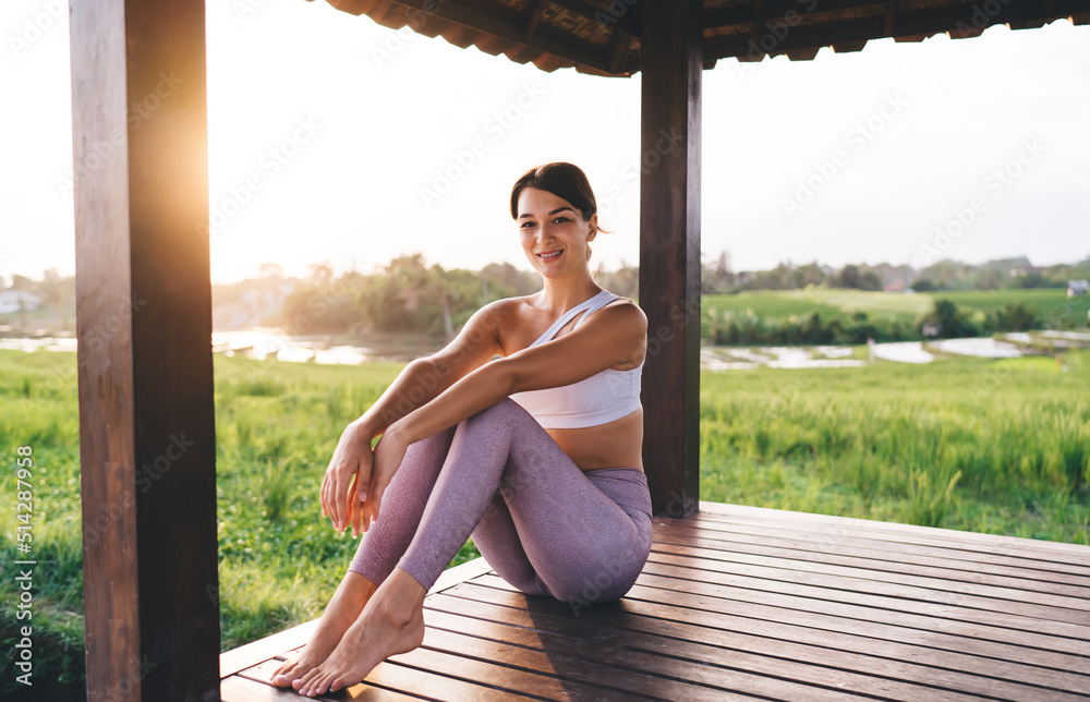 Portrait of happy Caucasian woman in sportive wear smiling at camera during morning sunshine in Thailand spending time for recreating and meditate, carefree fit girl in tracksuit posing at terrace