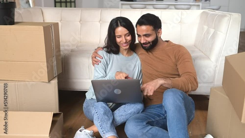 Lovely Indian couple choosing new furniture online in new house sitting on the floor in empty living room among cardboard boxes, multiracial man and woman with laptop at moving day