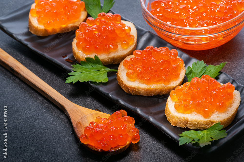Red expensive caviar on wooden spoon, on black rectangular plate on butter buns decorated with parsley, in glass bowl. Expensive caviar on spoon on buns and in glass plate. Red expensive caviar on wooden spoon, on black rectangular plate on butter buns decorated with parsley, in glass bowl. Expensive caviar on spoon on buns and in glass plate.