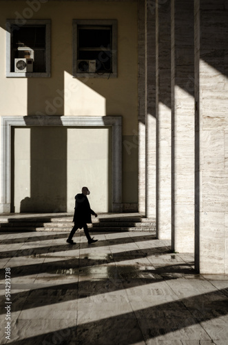 Silhouette of a person wearing a mask, who walks through a corridor surrounded by columns while sunlight filters through them