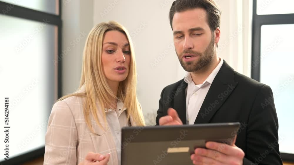Beautiful woman and serious man with a digital tablet in hands is standing at the office, watching at tablet screen and feeling confused Beautiful woman and serious man with a digital tablet in hands is standing at the office, watching at tablet screen and feeling confused