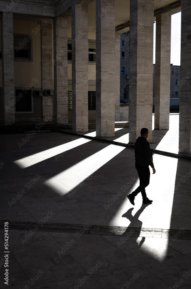 Sunlight filters through several columns of a building while a young man walks between them, revealing only his silhouette.