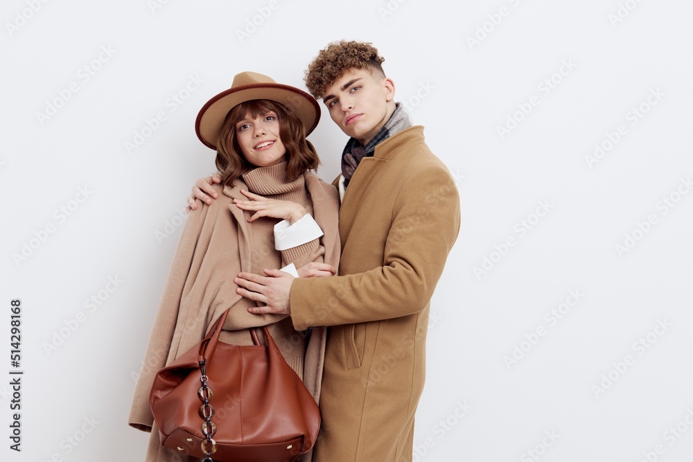 a man and a woman in an autumn coat stand on a white background and gently embrace looking at the camera, a woman has a hat on her head and she holds a bag in her hand a man and a woman in an autumn coat stand on a white background and gently embrace looking at the camera, a woman has a hat on her head and she holds a bag in her hand