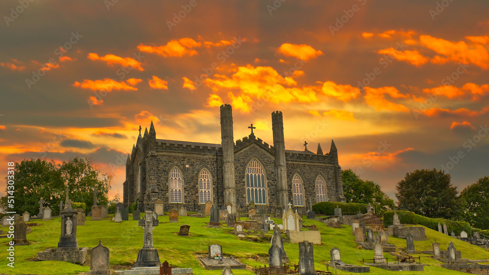 St. John RC Roman Catholic Chapel Churchyard Cemetery, Coleraine, Londonderry at the sunset with dramatic orange and yellow colour sky with clouds in the background and Tombstones in the foreground