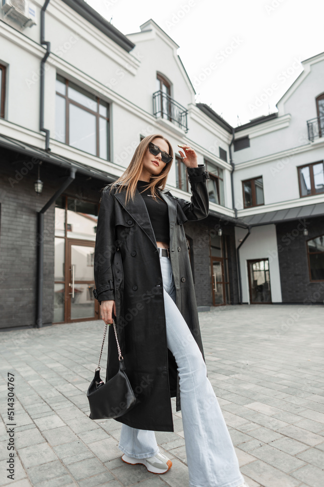 Fashionable beautiful model woman in cool black urban outfit with leather long  coat, jeans, trendy sunglasses and purse walking in the city Stock Photo |  Adobe Stock
