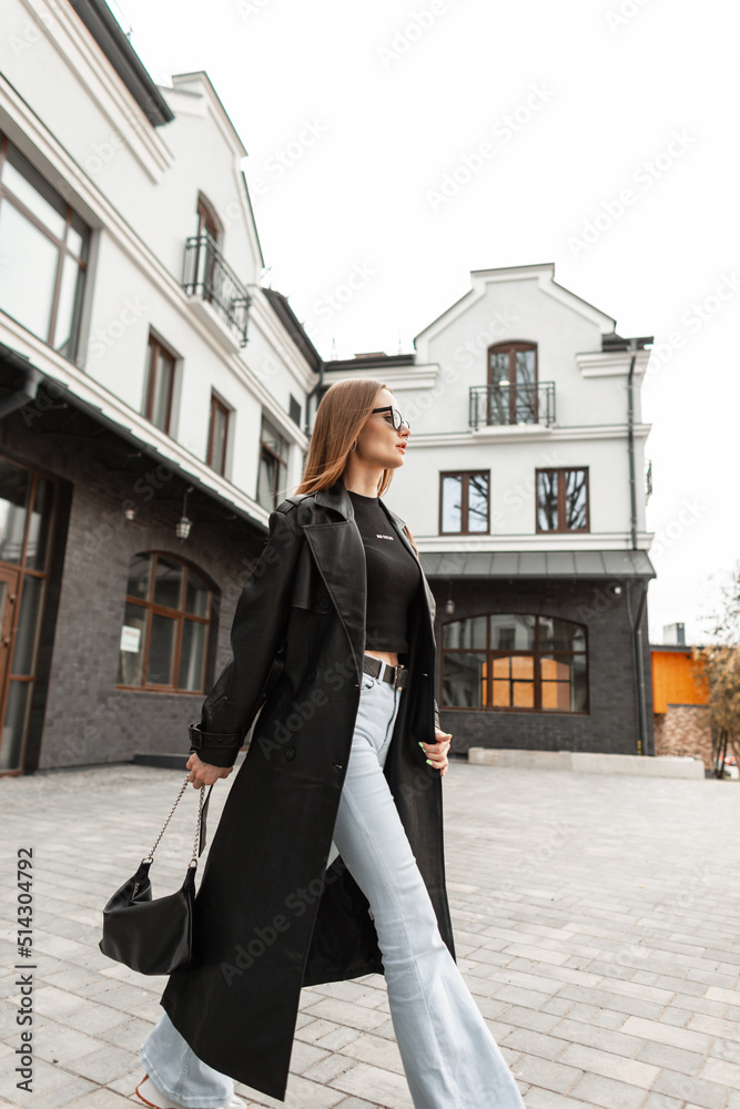 Fashion street beauty woman model with black fashionable outfit with long leather coat, jeans and stylish bag walks on the street