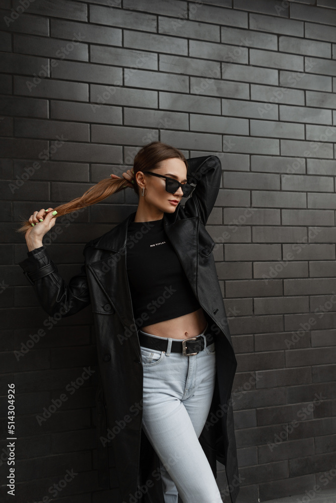 Fashion trendy beauty woman with fashionable eyewear in black stylish urban casual clothes with long coat, t-shirt and hight waist jeans stands and poses near a black brick background