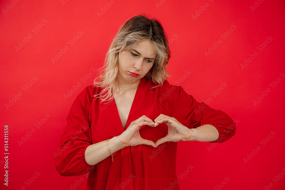 Portrait of upset beautiful young woman in holiday red look with perfect bright makeup isolated on red background. Unrequited love, making heart shape by hands. Fashion and beauty industry Portrait of upset beautiful young woman in holiday red look with perfect bright makeup isolated on red background. Unrequited love, making heart shape by hands. Fashion and beauty industry