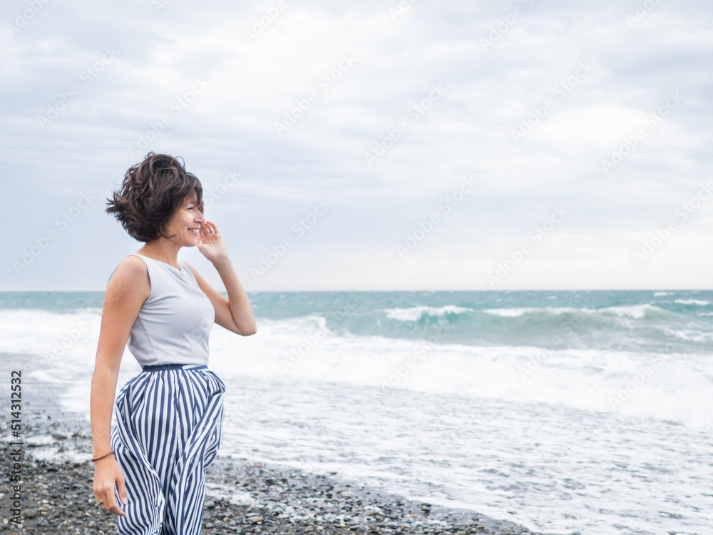Happy woman greets stormy sea on pebble beach. Woman with hair ruffled with the wind. Wanderlust concept. Vacation on sea coast. Happy woman greets stormy sea on pebble beach. Woman with hair ruffled with the wind. Wanderlust concept. Vacation on sea coast.