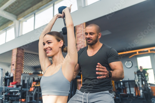 Athlete slim pretty girl lifting a dumbbell above her head being supported by a male good-looking fit personal trainer at the gym. High quality photo