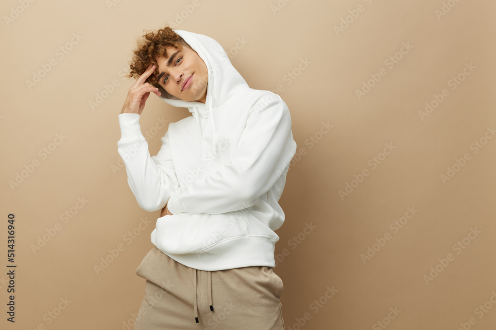 photo of a stylish young man in a white hoodie standing on a beige background in a relaxed pose with his hand near his hair photo of a stylish young man in a white hoodie standing on a beige background in a relaxed pose with his hand near his hair