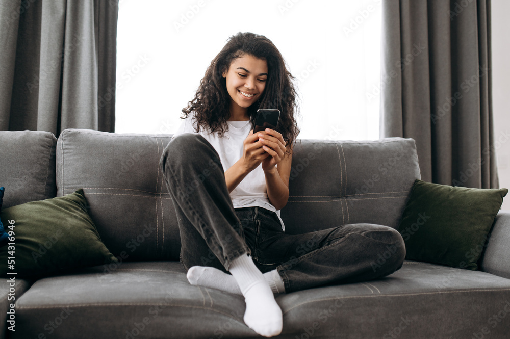 Modern girl blogging at social media, using smartphone. Pretty african american woman chatting with friends or family and smiling while sitting on sofa in living room, browsing internet, reading news Modern girl blogging at social media, using smartphone. Pretty african american woman chatting with friends or family and smiling while sitting on sofa in living room, browsing internet, reading news