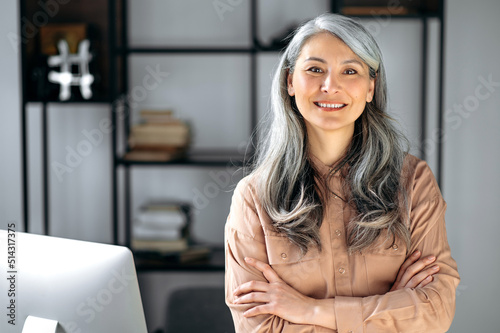 Portrait of a successful confident mature gray-haired lady, business woman, ceo or business tutor, standing in the office with arms crossed, looking and friendly smiling into the camera photo