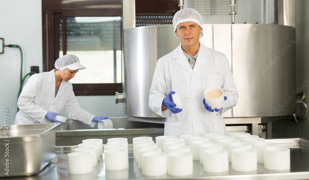 Portrait of professional cheesemaker wearing white uniform with cap and gloves working in shaping workshop of cheese factory