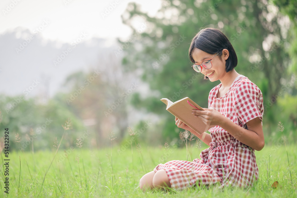 Cute asian teenager girl with glasses sitting on meadow reading book outdoors. Cute asian teenager girl with glasses sitting on meadow reading book outdoors.