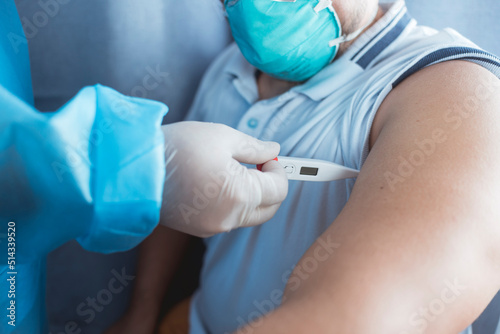 A nurse in PPE gear inserts a digital thermometer in the armpit of a man wearing a N95 mask. Measuring an underarm or axillary temperature. At the emergency ward of a hospital. photo