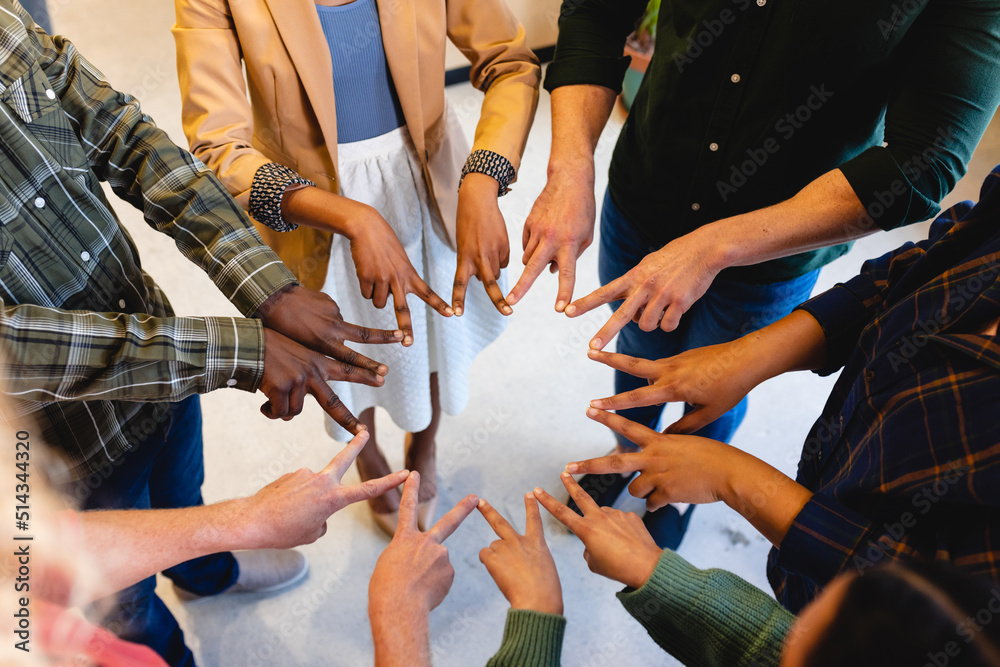 Multiracial male and female fashion designers huddling with peace signs in office Multiracial male and female fashion designers huddling with peace signs in office