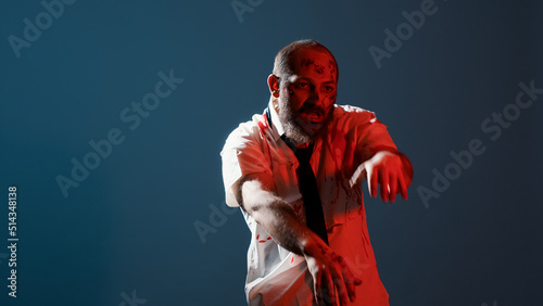 Creepy zombie growling at camera while standing on blue background. Dangerous and evil looking brain-eating monster with deep and bloody wounds moving hands while acting bizarre. Studio shot