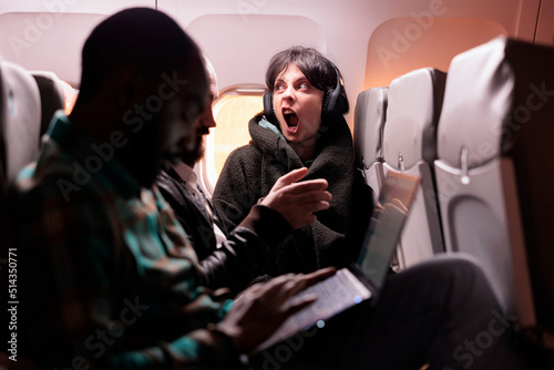 Multiethnic group of people flying abroad in economy class, using laptop, smartphone and headphones during sunset flight. Passengers travelling with international commercial airline.