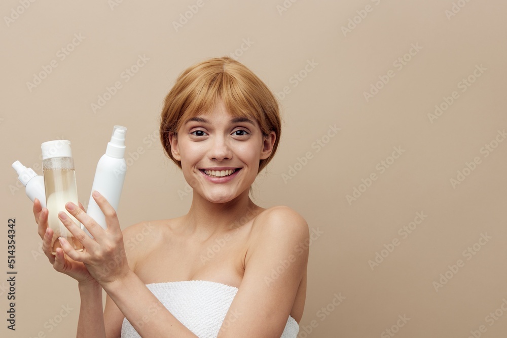 an attractive woman with short hair, wrapped in a towel, stands on a beige background holding various jars of care cosmetics in her hands, smiling sweetly an attractive woman with short hair, wrapped in a towel, stands on a beige background holding various jars of care cosmetics in her hands, smiling sweetly