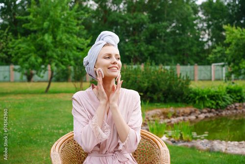 Smiling beautiful young woman with towel on head hold cotton pad cleansing face skin with cleaner, happy lady, remove makeup, enjoy healthy clean skin care treatment concept sitting in garden at home