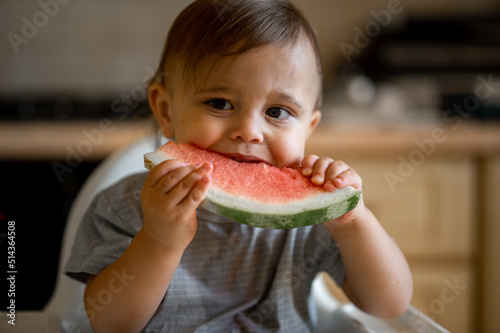 a one-year-old baby is sitting at home in the kitchen on a high chair and eating a watermelon