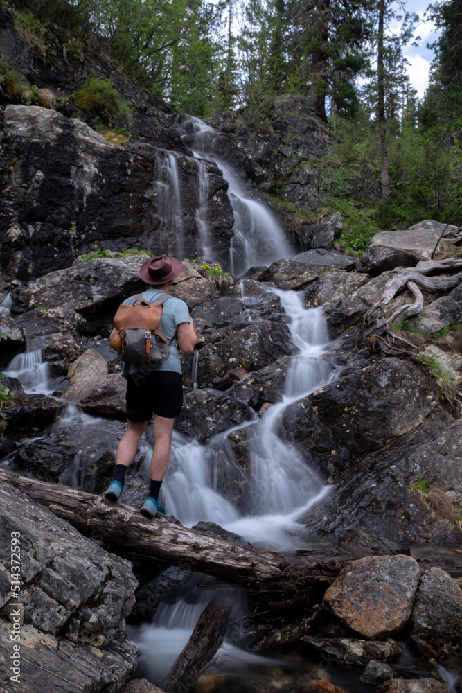 Tourist in hat with backpack walks along log against backdrop of waterfall Tourist in hat with backpack walks along log against backdrop of waterfall