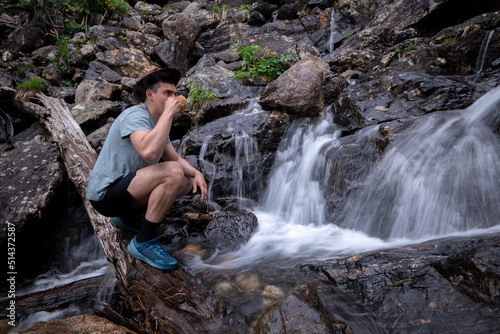 Young tourist in hat drinks from kuksa from waterfall in mountains