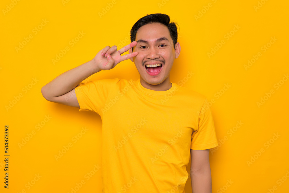 Cheerful young Asian man in casual t-shirt showing peace sign with fingers over face isolated on yellow background. People lifestyle concept Cheerful young Asian man in casual t-shirt showing peace sign with fingers over face isolated on yellow background. People lifestyle concept