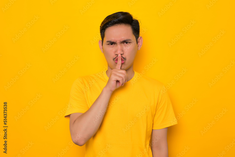 Attractive young Asian man in casual t-shirt showing shh gesture, press finger to lips isolated on yellow background. People lifestyle concept Attractive young Asian man in casual t-shirt showing shh gesture, press finger to lips isolated on yellow background. People lifestyle concept