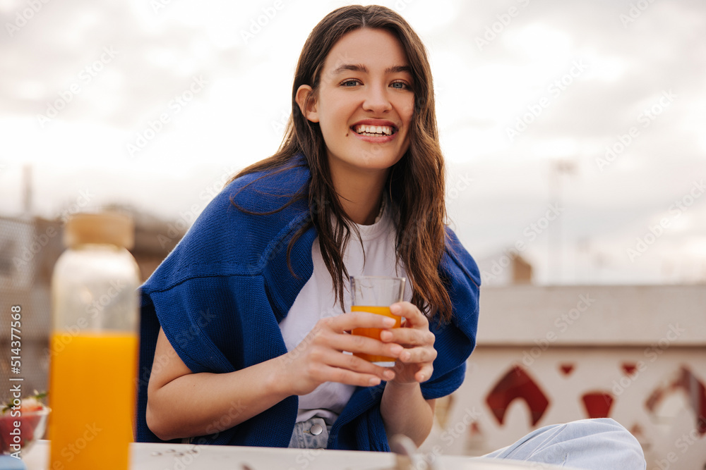 Happy young caucasian woman sitting on terrace in cafe on street of modern city. Brunette wearing hoodie and T-shirt drinking juice. Light breakfast concept Happy young caucasian woman sitting on terrace in cafe on street of modern city. Brunette wearing hoodie and T-shirt drinking juice. Light breakfast concept