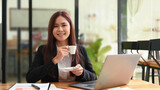 Positive young entrepreneur sitting front of laptop with coffee cup and smiling at camera