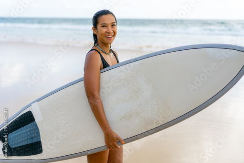 Photography Portrait of Young Asian woman surfer in swimwear holding surfboard walking on tropical beach at summer sunset