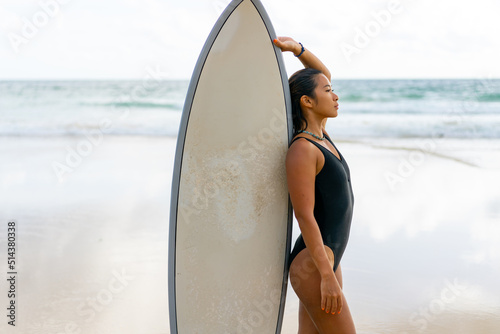 Photography Portrait of Young Asian woman surfer in swimwear holding surfboard walking on tropical beach at summer sunset