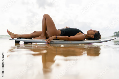 Canvas Print Beautiful Asian woman surfer in swimwear relaxing on surfboard after surfing at tropical beach at summer sunset