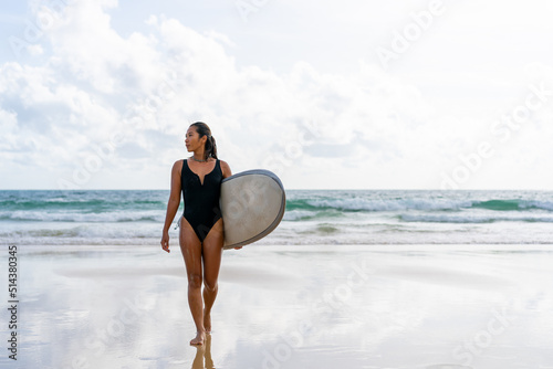 Canvas Print Portrait of Young Asian woman surfer in swimwear holding surfboard walking on tropical beach at summer sunset