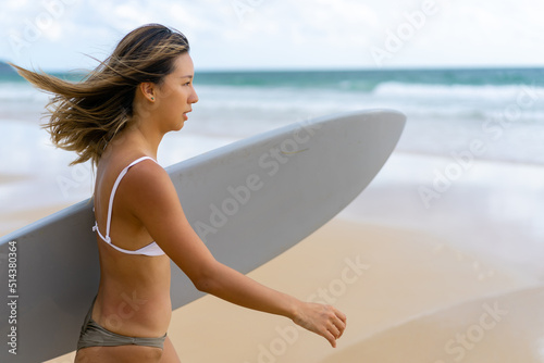 Photography Portrait of Young Asian woman surfer in swimwear holding surfboard walking on tropical beach at summer sunset