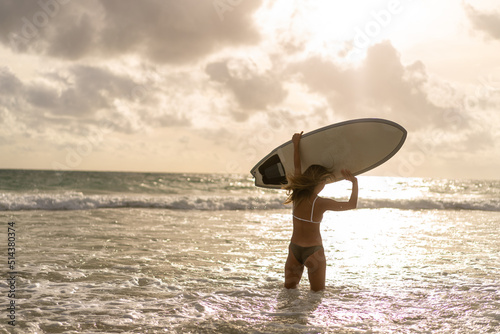 Portrait of Young Asian woman surfer in swimwear holding surfboard walking on tropical beach at summer sunset. Beautiful female enjoy outdoor activity lifestyle water sport surfing on summer vacation