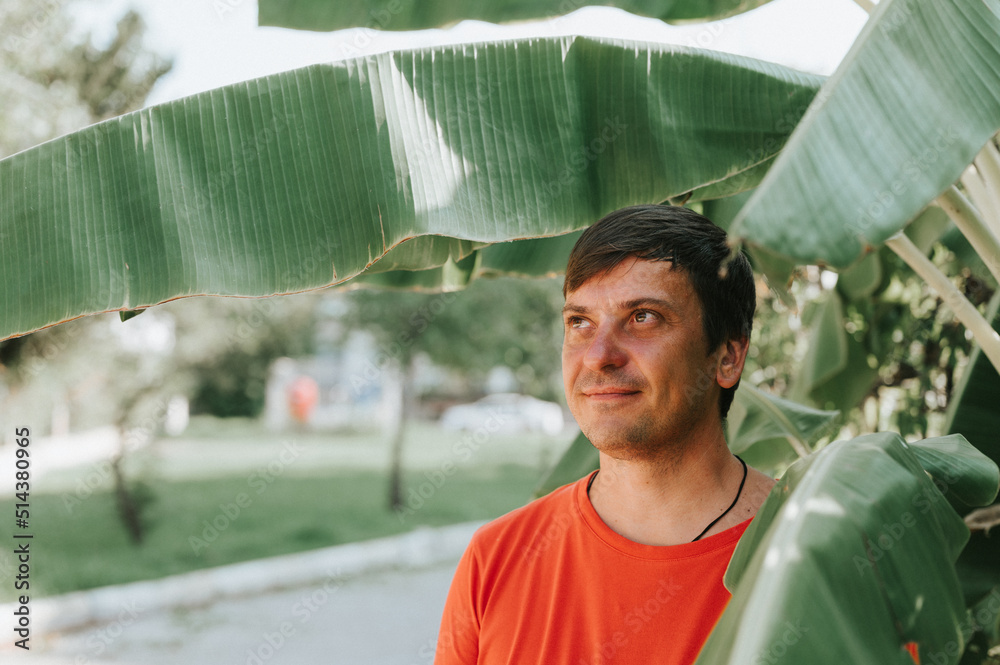 portrait of face happy smiling unshaven mature 40+ years man solo traveler with stubble on the background of green plants palm leaves in nature on sunny summer day on vacation travel getaway