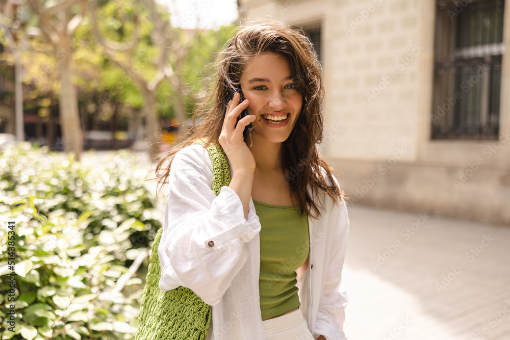 Beautiful young caucasian woman in good mood talking on phone looking at camera. Brunette wears casual clothes walking around city. Technology concept Beautiful young caucasian woman in good mood talking on phone looking at camera. Brunette wears casual clothes walking around city. Technology concept