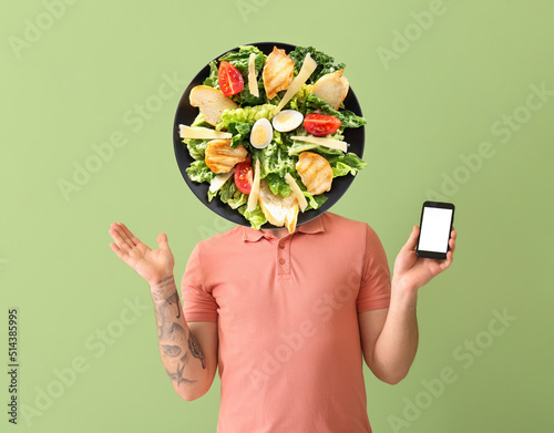 Young tattooed man with phone and plate of tasty Caesar salad instead of his head on green background photo