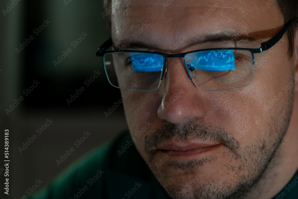 Portrait of millennial concentrated trader man in glasses looks at screen with graphs in dark. Caucasian man monitors exchange rate. Business Graph and chart of currency reflection in eyeglasses. Portrait of millennial concentrated trader man in glasses looks at screen with graphs in dark. Caucasian man monitors exchange rate. Business Graph and chart of currency reflection in eyeglasses.