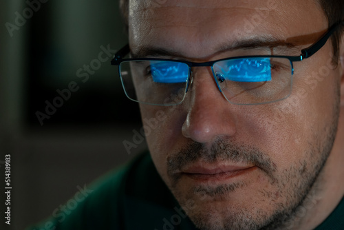 Portrait of millennial concentrated trader man in glasses looks at screen with graphs in dark. Caucasian man monitors exchange rate. Business Graph and chart of currency reflection in eyeglasses.