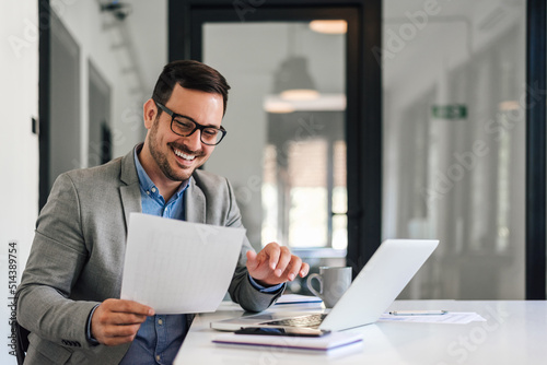 Smiling male executive reading documents while working on laptop at office desk