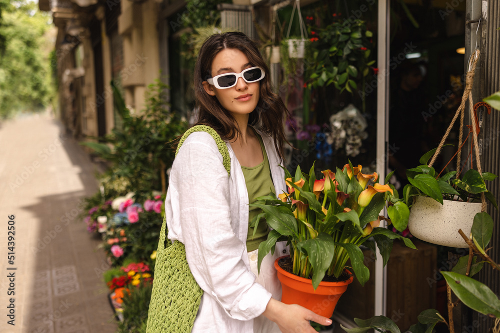 Calm young caucasian female shopper holding pot of flowers standing in market. Brunette wears casual clothes, sunglasses. Relaxed lifestyle, concept