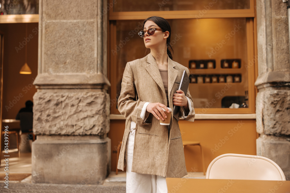 Fashionable young caucasian woman holding laptop, coffee standing outdoors near cafe. Brunette wears beige long jacket, sunglasses. City life concept