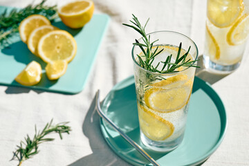 Summer refreshing lemonade drink or alcoholic cocktail with ice, rosemary and lemon slices on the table in the garden. Fresh healthy cold lemon beverage. Water with lemon.