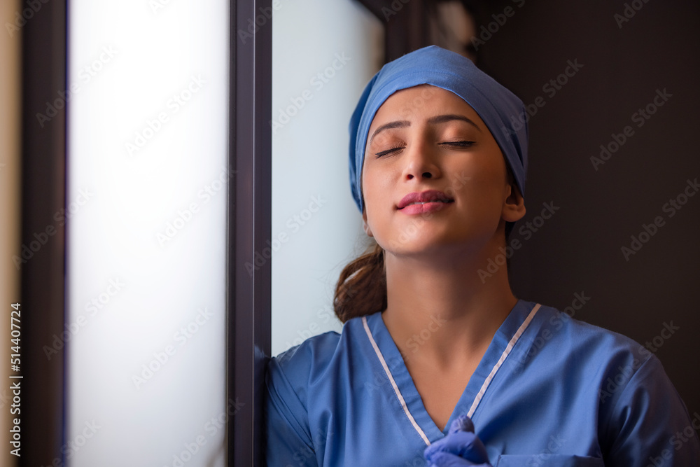 Close-up of a exhausted female nurse standing by window with her eyes closed in hospital. Close-up of a exhausted female nurse standing by window with her eyes closed in hospital.