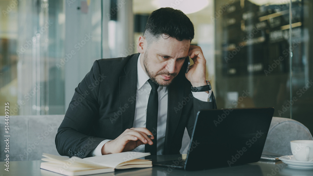 Smart Caucasian businessman sitting, talking on smartphone and writing down business information in his notepad while looking at laptop in glassy cafe Smart Caucasian businessman sitting, talking on smartphone and writing down business information in his notepad while looking at laptop in glassy cafe